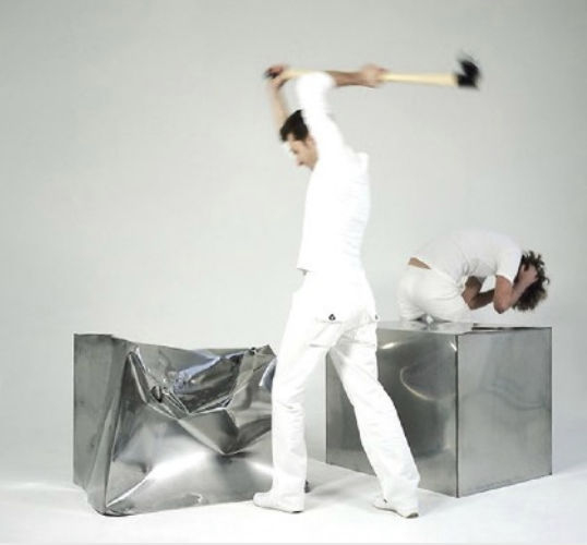 Zwei Männer in Weiß, einer schlägt mit Hammer auf eine Metallkiste, der andere kauert auf einer zweiten Kiste.